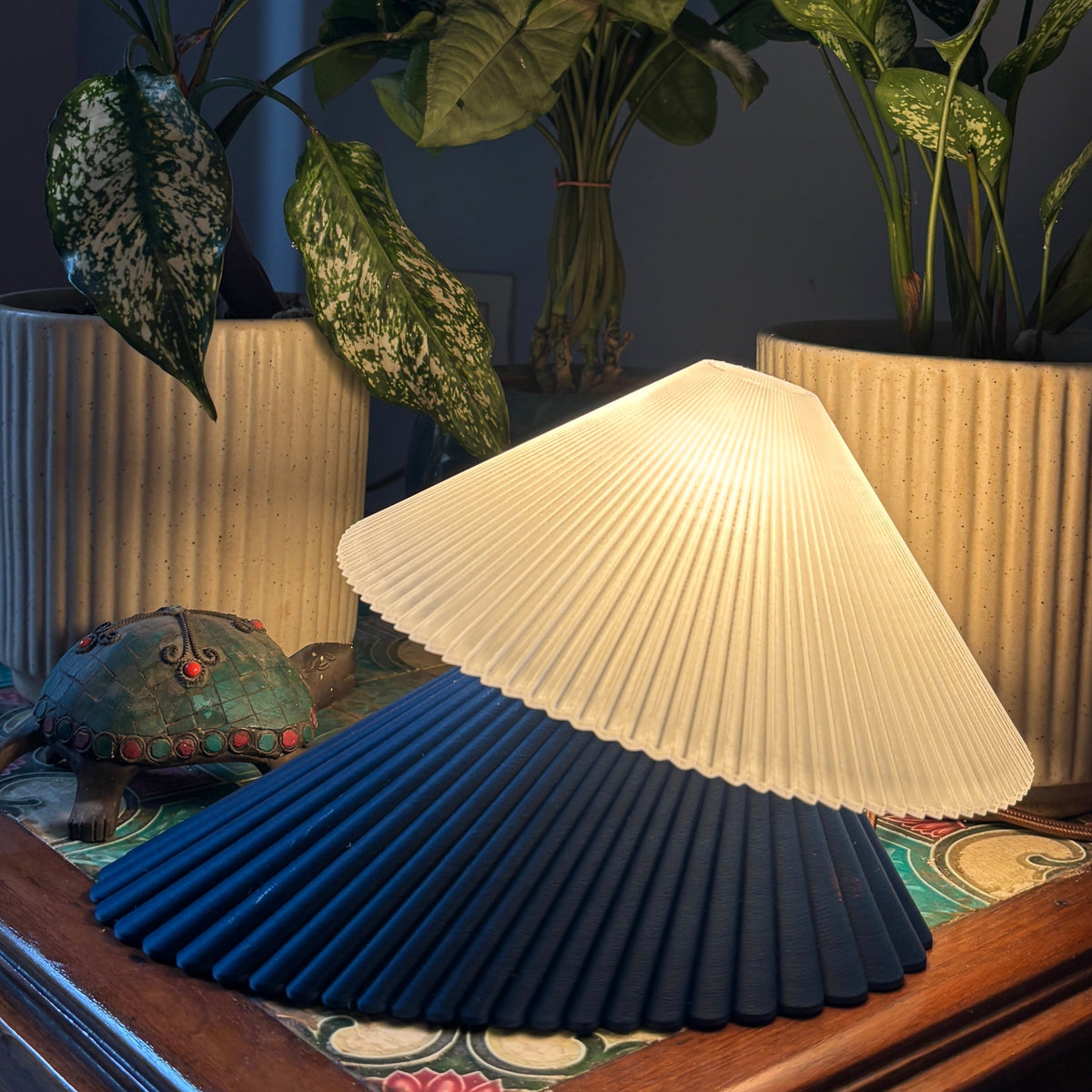 A pleated table lamp with a dark navy blue ribbed base and a glowing warm white pleated shade, styled on a wooden table between potted green plants in a dimly lit room.